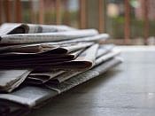 A stack of newspapers sitting on the left side of a desk.