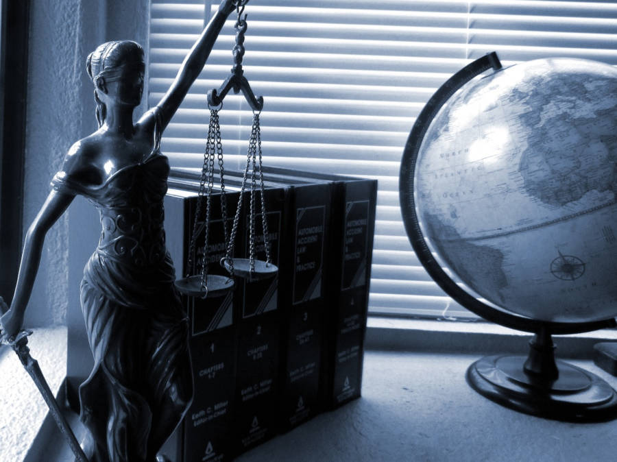 Lady Justice sitting on a desk with a background of books and a globe.