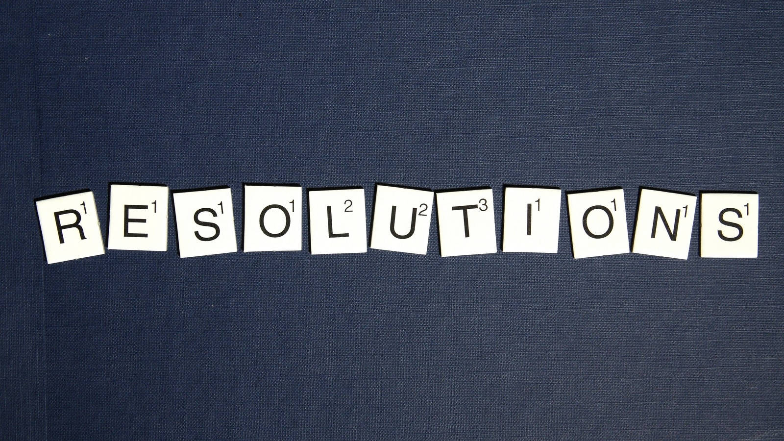 Scrabble tile pieces spelling "resolutions" set atop a blue fabric background.